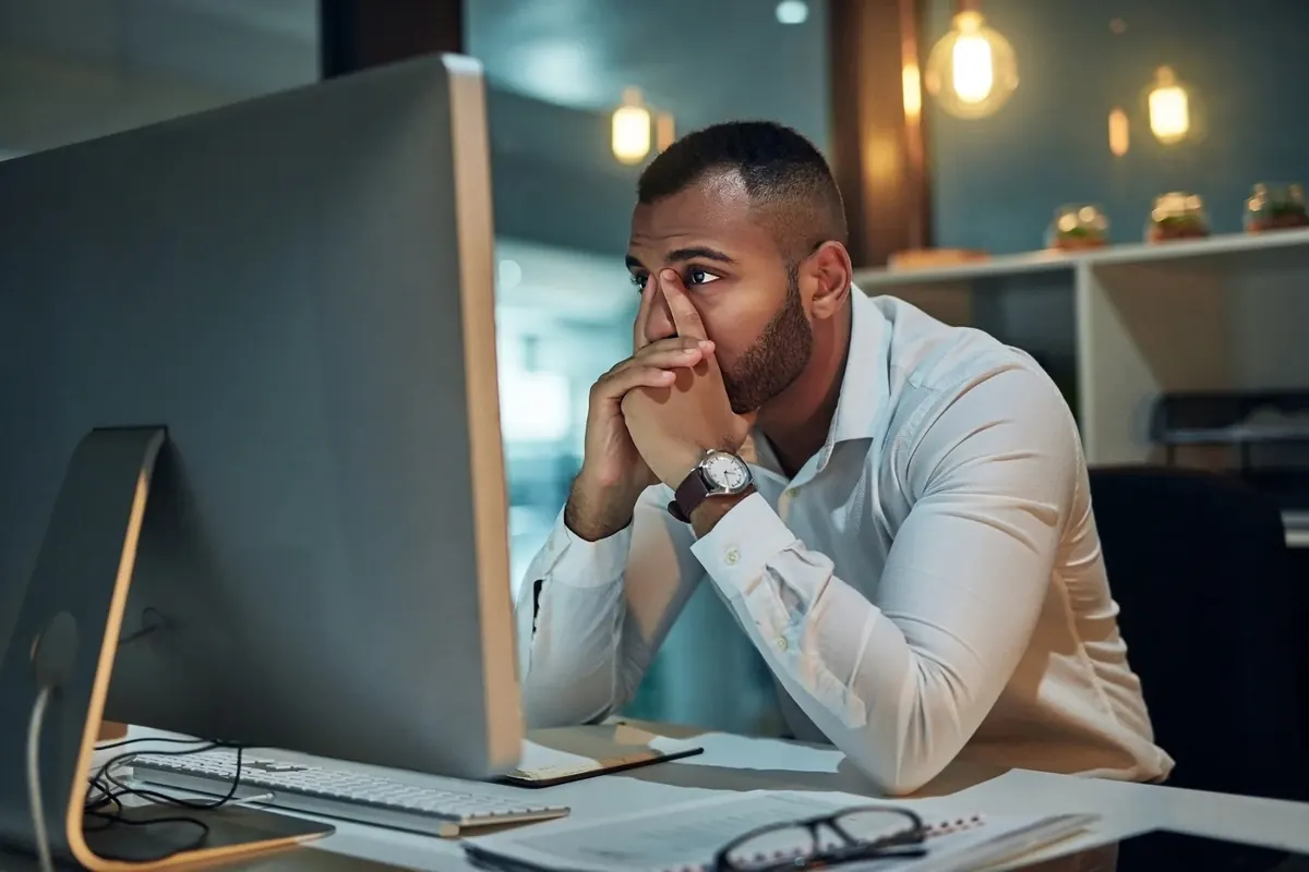 A man at an office desk in the evening, hands to his face, looking at a computer screen